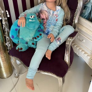 May include: A doll wearing light blue pajamas with a graphic of Elsa from Frozen. The doll is seated on a purple velvet chair and holding a teal plush monster toy with purple spots. The chair has silver painted trim.