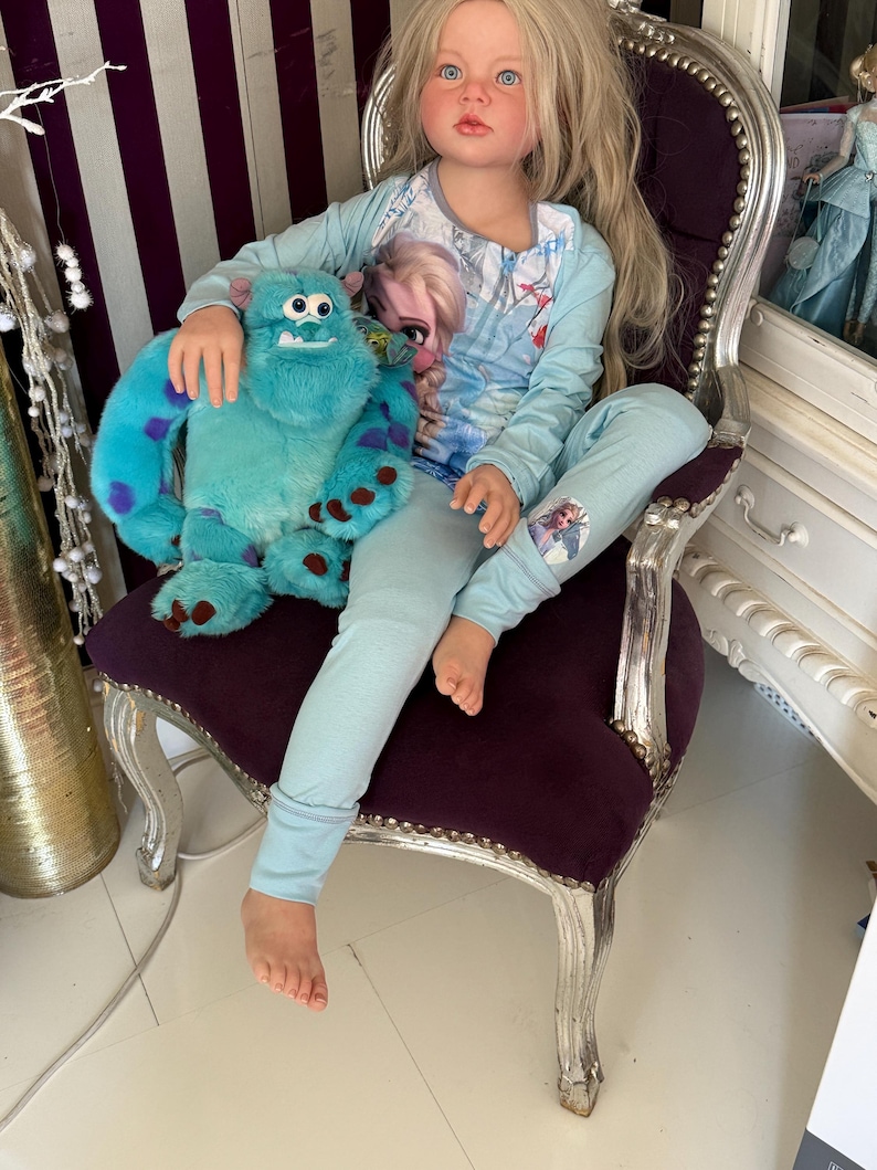 May include: A doll wearing light blue pajamas with a graphic of Elsa from Frozen. The doll is seated on a purple velvet chair and holding a teal plush monster toy with purple spots. The chair has silver painted trim.