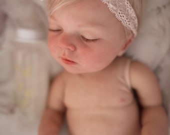 May include: A realistic newborn doll with closed eyes and fair skin, wearing a pale pink lace headband adorned with a matching fabric flower. The doll has light blonde hair and is positioned in a resting pose, with a blurred baby bottle nearby.