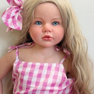 May include: A lifelike doll with long blonde hair and bright blue eyes. The doll wears a pink and white gingham dress with matching bow. The dress has tie straps. The doll's skin has a realistic tone.