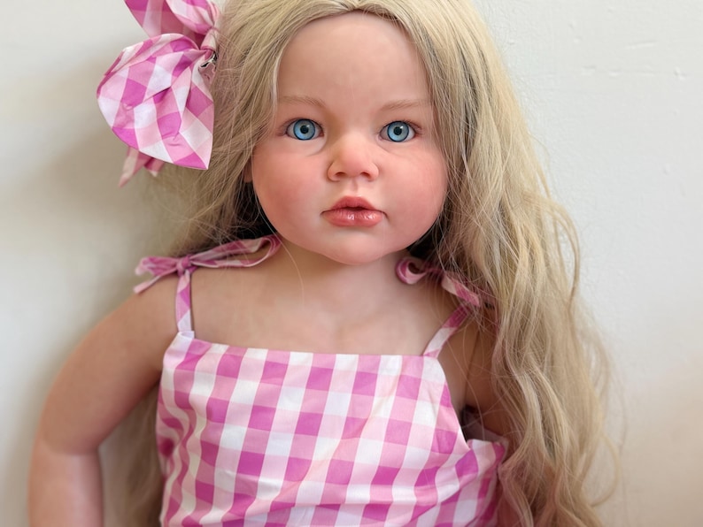May include: A lifelike doll with long blonde hair and bright blue eyes. The doll wears a pink and white gingham dress with matching bow. The dress has tie straps. The doll's skin has a realistic tone.