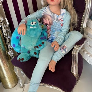 May include: A doll wearing light blue pajamas with a graphic of Elsa from Frozen. The doll is holding a blue stuffed animal with purple spots. The doll is sitting in a purple chair with silver trim.