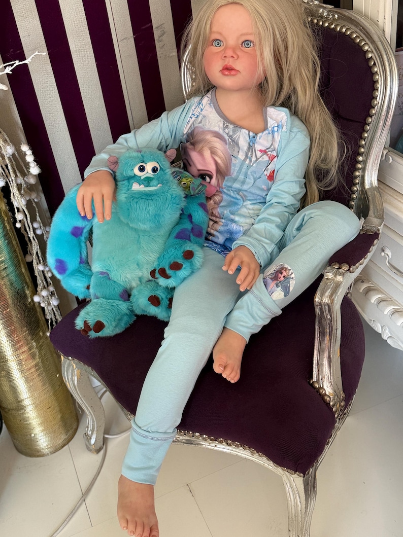 May include: A doll wearing light blue pajamas with a graphic of Elsa from Frozen. The doll is holding a blue stuffed animal with purple spots. The doll is sitting in a purple chair with silver trim.