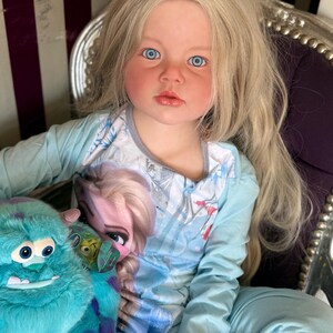 May include: A young child with long blonde hair and blue eyes wearing a light blue long-sleeved shirt and pants. The shirt features a graphic of Elsa from Frozen. The child is holding a teal plush monster toy.