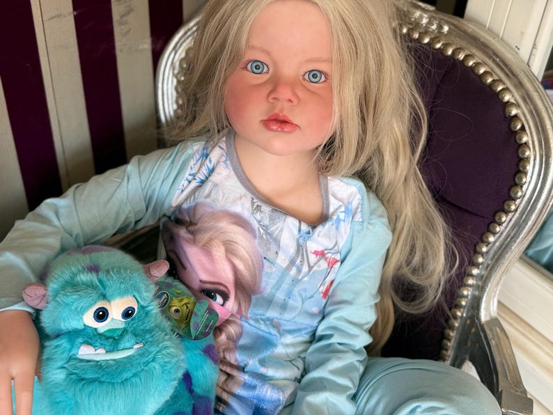 May include: A young child with long blonde hair and blue eyes wearing a light blue long-sleeved shirt and pants. The shirt features a graphic of Elsa from Frozen. The child is holding a teal plush monster toy.