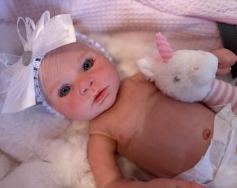 May include: A lifelike baby doll with fair skin, blue eyes, and a white headband with a bow. The doll is holding a small white unicorn plush toy with a pink horn. The doll is wearing a diaper and lying on a fluffy white surface.