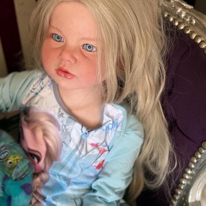 May include: A doll with long blonde hair and blue eyes is seated in a decorative chair. The doll is wearing a light blue long-sleeved shirt with a graphic of a princess. A blue and green stuffed animal is in the doll's lap.