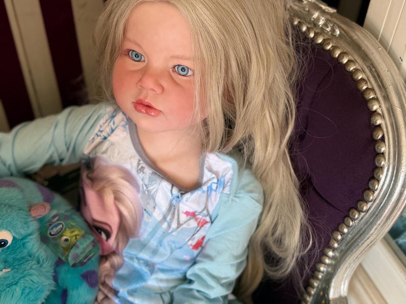 May include: A doll with long blonde hair and blue eyes is seated in a decorative chair. The doll is wearing a light blue long-sleeved shirt with a graphic of a princess. A blue and green stuffed animal is in the doll's lap.