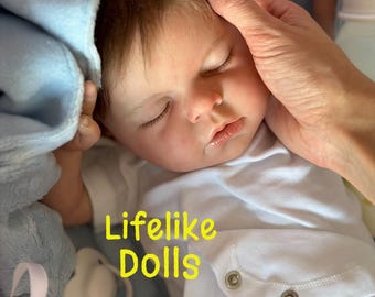 May include: A realistic baby doll with brown hair and closed eyes, wearing a white short-sleeved bodysuit with two metal snaps at the bottom. The doll is wrapped in a light blue blanket and positioned next to a white pacifier. Text reads Lifelike Dolls.
