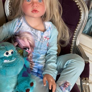 May include: A doll with long blonde hair and blue eyes wearing a light blue pajama set with a Frozen theme. The doll is holding a teal plush monster toy. The doll is seated in a purple chair with silver trim.
