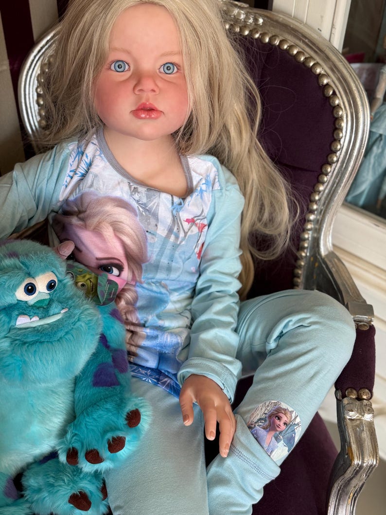 May include: A doll with long blonde hair and blue eyes wearing a light blue pajama set with a Frozen theme. The doll is holding a teal plush monster toy. The doll is seated in a purple chair with silver trim.