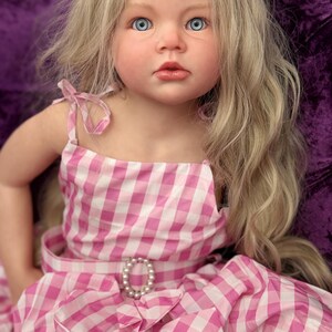 May include: A lifelike doll with blue eyes and long blonde hair. The doll wears a pink and white gingham dress with a pearl-adorned belt buckle. The dress has a bow detail at the waist and shoulder straps.
