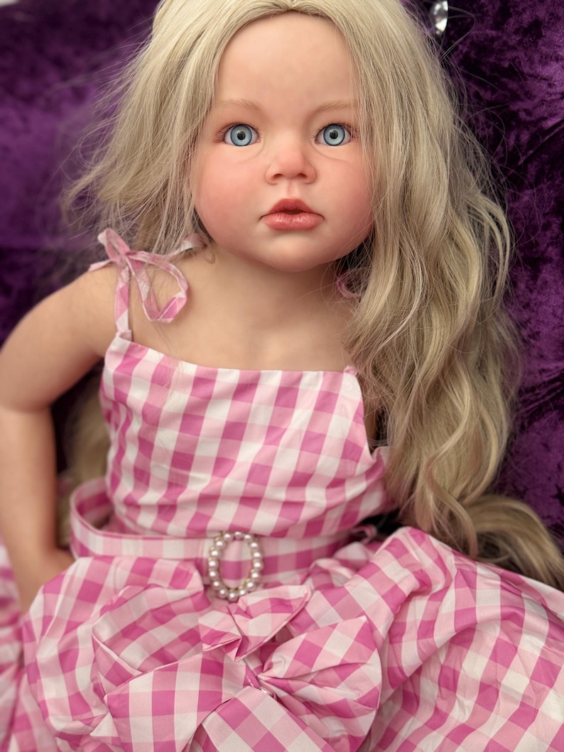 May include: A lifelike doll with blue eyes and long blonde hair. The doll wears a pink and white gingham dress with a pearl-adorned belt buckle. The dress has a bow detail at the waist and shoulder straps.