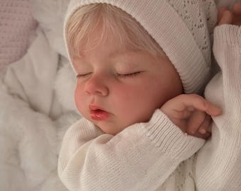 May include: A realistic baby doll with pale skin and blonde hair, wearing a white knitted hat with a small bow and a matching white long-sleeved sweater featuring a delicate openwork pattern. The doll has closed eyes and a soft, sleeping expression.