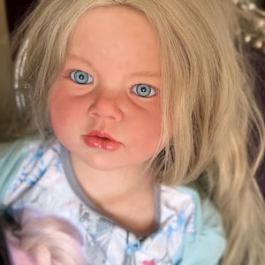 May include: A lifelike doll with fair skin, rosy cheeks, and light blonde hair. The doll has bright blue eyes and a slightly open mouth. It is wearing a light blue and white patterned outfit. The doll's hair is long and wavy, framing its face.