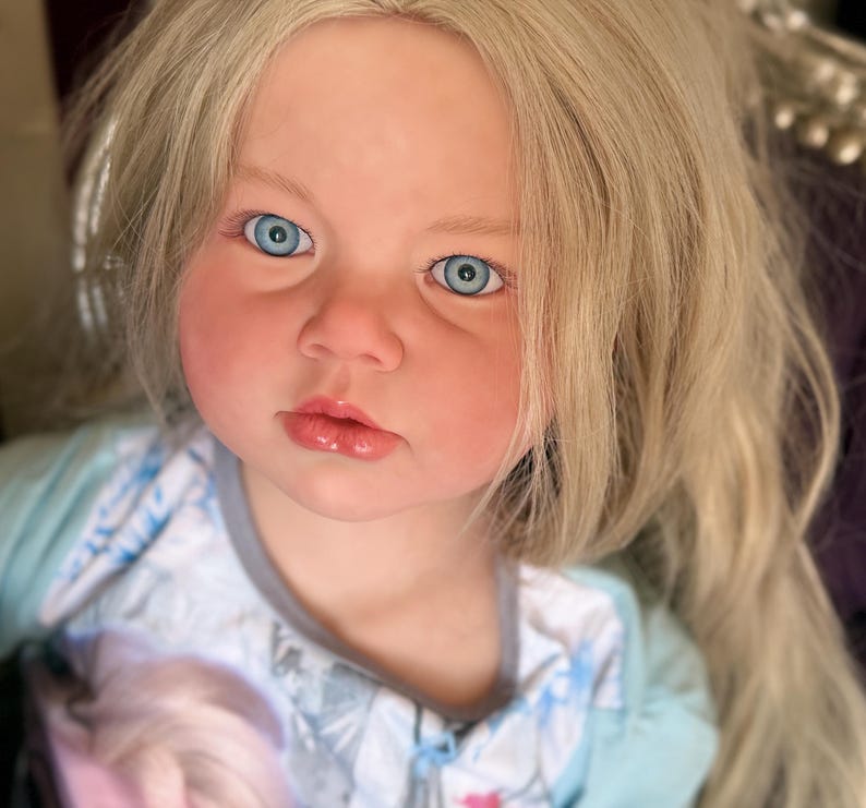 May include: A lifelike doll with fair skin, rosy cheeks, and light blonde hair. The doll has bright blue eyes and a slightly open mouth. It is wearing a light blue and white patterned outfit. The doll's hair is long and wavy, framing its face.