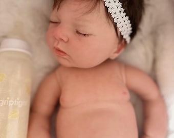 May include: A realistic sleeping baby doll with dark brown hair and a white lace headband featuring a small bow. The doll has light skin, closed eyes, and is wearing a white diaper. Beside the doll is a clear plastic bottle with yellow leaf prints.