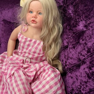 May include: A lifelike doll with blonde hair and blue eyes, wearing a pink and white gingham dress with a bow and a pearl-accented belt buckle. The doll is seated on a plush, deep purple velvet surface.