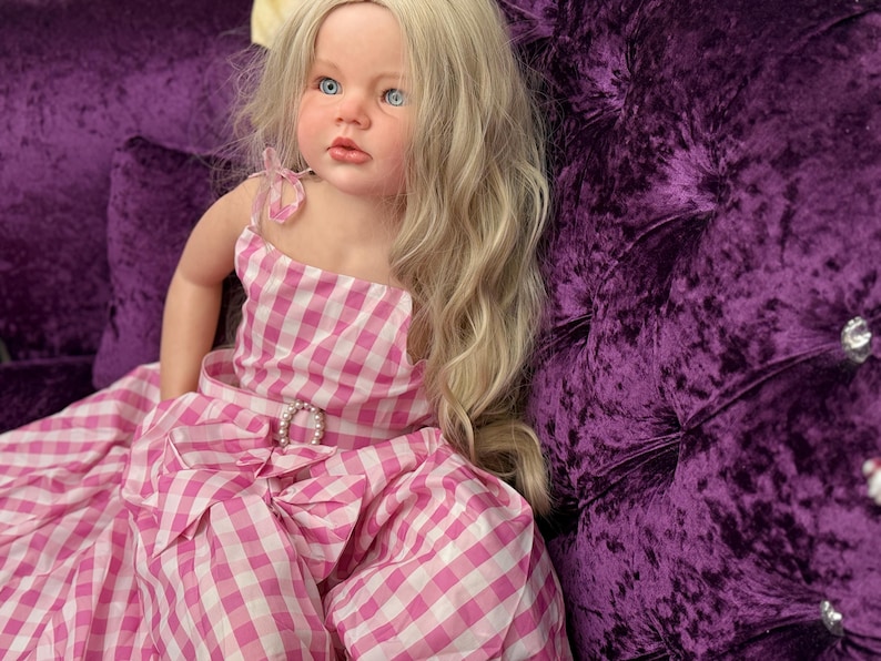 May include: A lifelike doll with blonde hair and blue eyes, wearing a pink and white gingham dress with a bow and a pearl-accented belt buckle. The doll is seated on a plush, deep purple velvet surface.