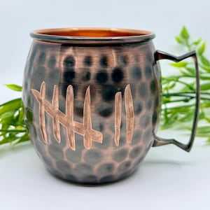 May include: A copper-colored Moscow Mule mug with a hammered finish and a handle. The mug features etched tally marks on the front. The interior of the mug is a lighter copper color.