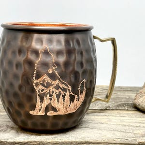 May include: A hammered copper mug with a brass handle features an etched design of a wolf howling at the moon in front of a mountain range and pine trees.