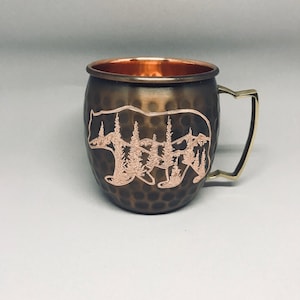 May include: A hammered copper mug with a bear and trees design. The mug has a brass handle and a copper interior.