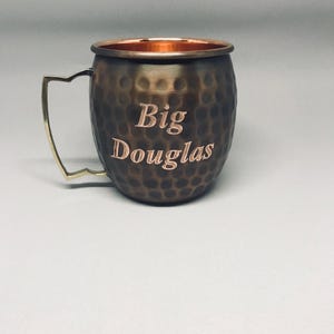 May include: A hammered copper mug with a brass handle. The mug has the words "Big Douglas" engraved on the side.