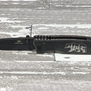 May include: Black folding pocket knife with a bear and trees design engraved on the handle. The knife has a black blade and a silver "Buck Knives" logo on the blade.