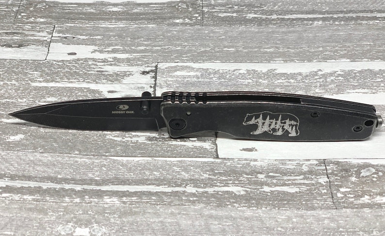 May include: Black folding pocket knife with a bear and trees design engraved on the handle. The knife has a black blade and a silver "Buck Knives" logo on the blade.