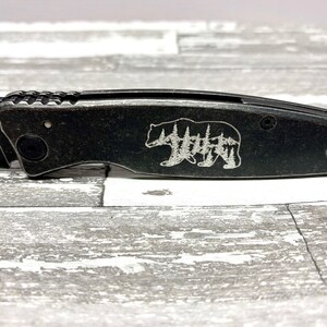 May include: A black pocket knife with a bear and trees design engraved on the handle.