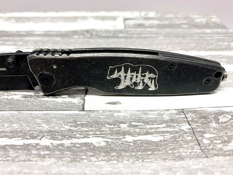 May include: A black pocket knife with a bear and trees design engraved on the handle.