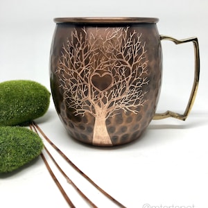 May include: A copper mug with a hammered finish and a tree of life design with a heart at the center. The mug has a brass handle.