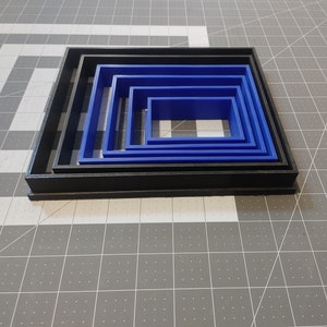 May include: A set of five nesting square cookie cutters. The largest cutter is black and the four smaller cutters are blue.