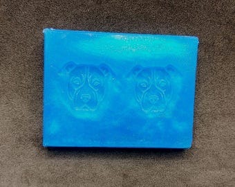 May include: A rectangular, blue soap bar with two embossed dog faces. The soap has a marbled appearance with lighter blue and white swirls. The dog faces are detailed with visible eyes, noses, and ears. The soap is a solid block.