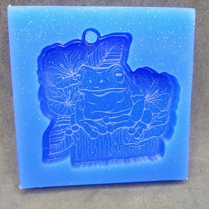 May include: Blue silicone mold with a detailed design of a frog sitting on a log surrounded by leaves and flowers.