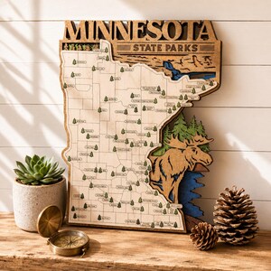 Könnte beinhalten: Eine lasergeschnittene Holzkarte der Minnesota State Parks mit eingravierten Parknamen, kleinen grünen Baum-Symbolen und einem großen Elch-Ausschnitt vor blauem Wasser und grünen Kiefern, mit dem Text MINNESOTA STATE PARKS oben.