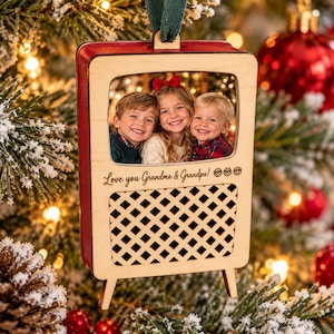 Op de afbeelding: Een houten kerstboomornament in de vorm van een vintage televisie met een foto van drie kinderen erin. Het frame bevat de gegraveerde tekst "Love you Grandma & Grandpa!" en een ruitpatroon aan de onderzijde.