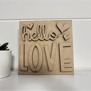 以下が含まれることがあります： 「hello LOVE」の文字が切り抜かれた木製のサイン。サインには3つのハートの形があり、明るい茶色の木材で作られています。サインの左側には、緑の植物が入った小さな白い鉢があります。