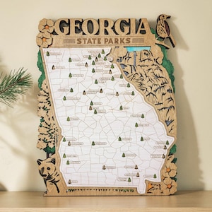 Peut inclure: Carte en bois des parcs d'État de Géorgie, représentant le contour de l'État avec les emplacements des parcs marqués par de petits arbres. La carte comprend le texte "GEORGIA STATE PARKS" en haut, avec des éléments décoratifs d'un oiseau, d'un ours et de fleurs.