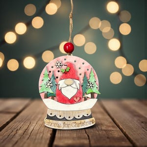 Op de afbeelding: Houten kerstornament in de vorm van een sneeuwbol, met een kabouter met een rode hoed en baard, omringd door groene bomen en sneeuwvlokken. De basis leest "Gnome for Christmas". Het ornament hangt aan een touwtje.