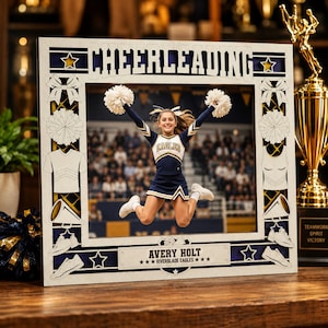 May include: A rectangular wooden picture frame featuring the word CHEERLEADING at the top and AVERY HOLT RIVERBLADE EAGLES at the bottom. The frame is decorated with cutouts of bows, pom-poms, uniforms, megaphones, and shoes, surrounding a photo.