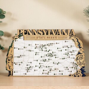 May include: Wooden map of Pennsylvania State Parks. The map is in the shape of the state, with the name "PENNSYLVANIA" at the top. The map has small green tree icons and is surrounded by a decorative border with a deer and fish.