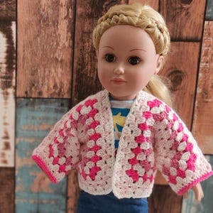 A pink and white crocheted cardigan with a loose fit, perfect for a doll. The cardigan has a simple open front and long sleeves.