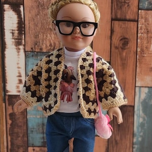 Pode incluir: Uma boneca vestindo uma jaqueta marrom e bege de croch&ecirc;, uma camiseta branca com um gr&aacute;fico e jeans azuis. A boneca usa &oacute;culos pretos e uma bolsa rosa. O fundo &eacute; um painel de madeira.