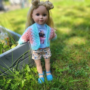 Pode incluir: Boneca com cabelo loiro em dois coques, vestindo uma camiseta azul clara com um desenho de pug, um cardig&atilde; de croch&ecirc; em tons pastel rosa, branco e azul, e shorts com estampa floral. A boneca tamb&eacute;m usa sand&aacute;lias azuis.