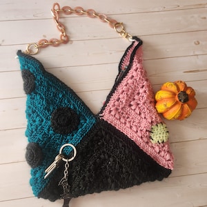 Peut inclure: Un sac crocheté à la main avec un design géométrique unique. Le sac présente des sections de fil bleu sarcelle, noir et rose, avec des accents circulaires noirs. Il a une bandoulière en chaîne rose et une petite décoration de citrouille.
