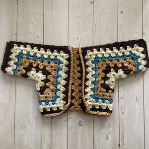 A crocheted cardigan with a dark brown, brown, cream, and blue color pattern. The cardigan is open in the front and has a loose fit.