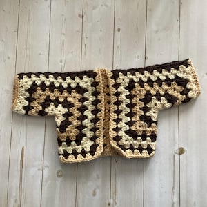 A crocheted cardigan with a dark brown, light brown and cream,  color pattern. The cardigan is open in the front and has a loose fit.