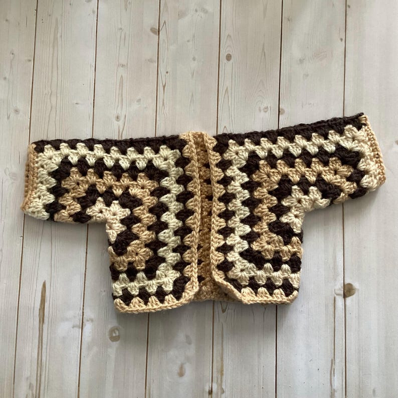 A crocheted cardigan with a dark brown, light brown and cream,  color pattern. The cardigan is open in the front and has a loose fit.