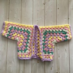 A crocheted cardigan with a white, pink, yellow, purple, pink, green, white, pink and yellow color pattern. The cardigan is open in the front and has a loose fit.